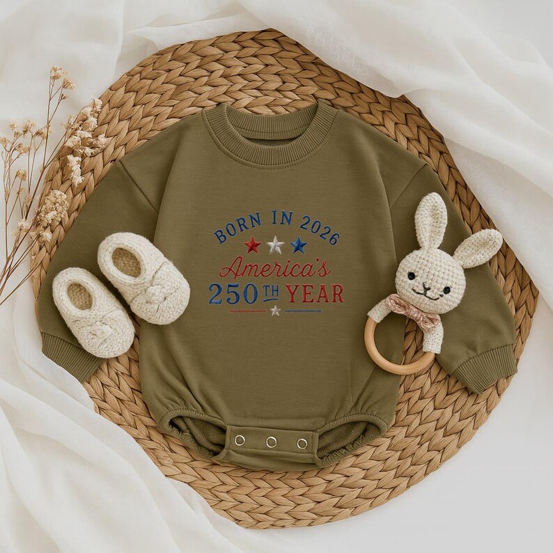 May include: Olive green baby bodysuit with the text "Born in 2026 America's 250th Year" embroidered in red, white, and blue. Includes a pair of cream-colored baby shoes and a bunny-shaped wooden teether.