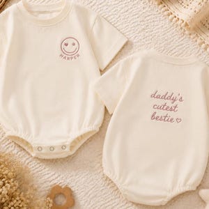Pode incluir: Dois macacões de bebê creme. Um tem uma carinha sorridente com uma estrela e um coração, e o nome "HARPER" bordado em roxo. O outro diz "daddy's cutest bestie" em escrita roxa com um coração.