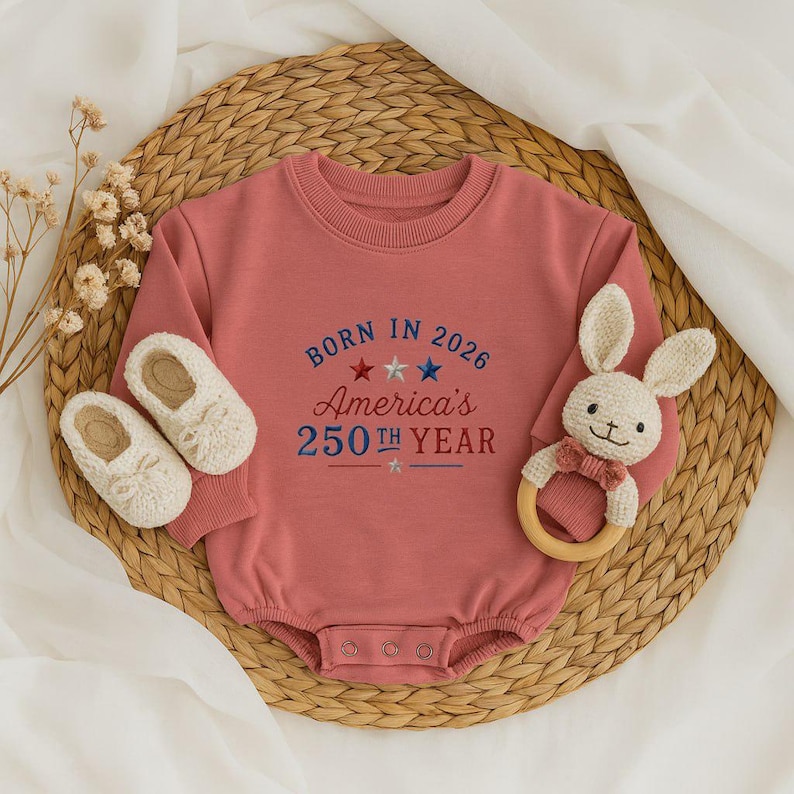 May include: A dusty rose baby onesie with the text "BORN IN 2026 America's 250th YEAR" in blue and red embroidery. The onesie is paired with white baby shoes and a bunny-shaped wooden teether.