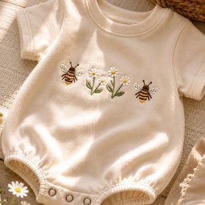 May include: A cream-coloured short-sleeve romper featuring an embroidered design of two bees flanking a pair of white daisies with green stems. The garment has a ribbed crew neckline, short sleeves, and a bottom closure with three metal snaps.