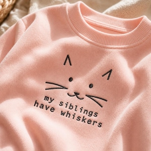 May include: A light pink crewneck sweatshirt featuring a ribbed collar and black embroidered cat face details. Below the cat face, the text "my siblings have whiskers" is embroidered in a simple, dark, sans-serif font on the chest area.