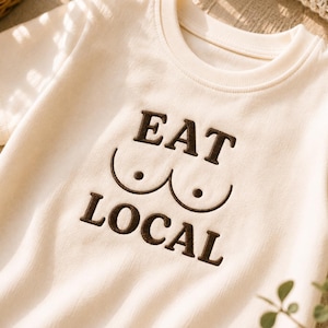 Puede incluir: Una camiseta de manga corta de color crema con bordado marrón oscuro en el pecho. El diseño consiste en el texto EAT sobre un gráfico de dos líneas curvas con puntos, y la palabra LOCAL debajo del gráfico, todo en una fuente negrita.