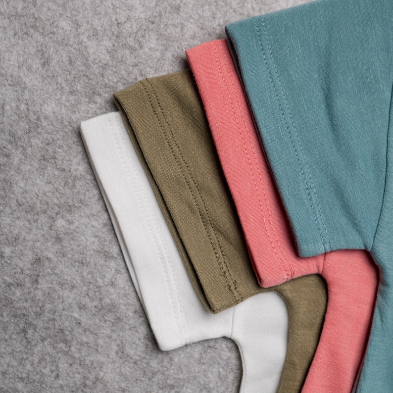 May include: A close-up of a stack of folded t-shirts in various colors. The colors include white, olive green, coral, and teal. The shirts are made of a soft material and have a simple design.