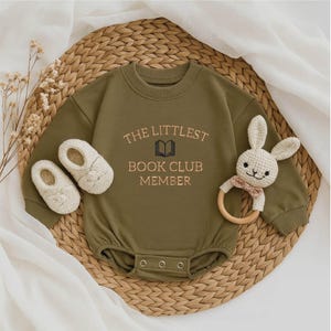 May include: An olive green baby romper with the embroidered text "THE LITTLEST BOOK CLUB MEMBER" in a light brown color. The image also includes a pair of cream-colored baby shoes and a bunny-shaped wooden teether.