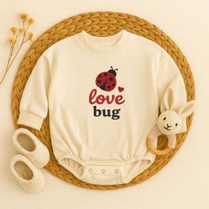 May include: Cream-colored baby onesie with a red and black ladybug design and the words "love bug." The onesie is paired with cream-colored baby shoes and a plush bunny rattle with a wooden ring.