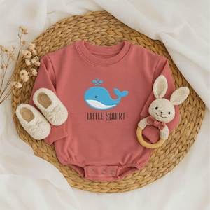 May include: A dusty rose baby romper with a blue whale design and the text "LITTLE SQUIRT". The image includes a pair of white baby shoes and a stuffed bunny with a wooden ring.