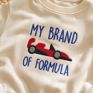 Könnte beinhalten: Ein cremefarbenes Sweatshirt mit geripptem Rundhalsausschnitt und blauem Sticktext, der MY BRAND OF FORMULA lautet. Zwischen den Textzeilen befindet sich ein gesticktes rotes und schwarzes Rennauto mit grauen Rädern.