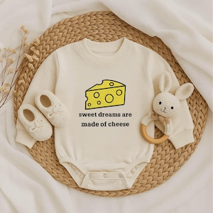 May include: Cream-colored baby onesie with a yellow cheese graphic and the text "sweet dreams are made of cheese." Includes matching baby shoes and a white bunny rattle with a wooden ring.