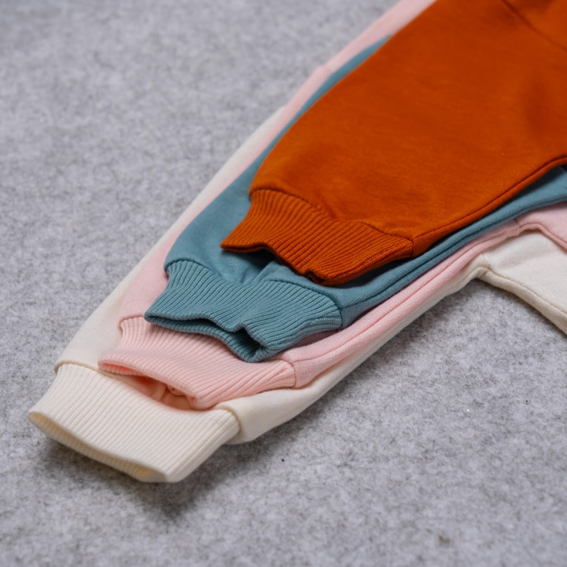 May include: A stack of four sweatshirts with ribbed cuffs in cream, pink, teal, and orange. The sleeves are layered, showcasing the different colors and textures of the fabric. The sweatshirts are likely made of a soft, comfortable material.