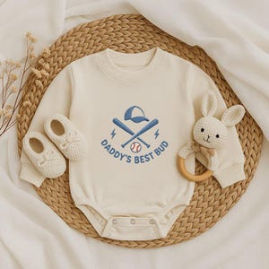 May include: Cream baby bodysuit with a baseball design, featuring crossed bats, a baseball, and a cap. The text "Daddy's Best Bud" is embroidered below. Includes matching baby shoes and a crochet bunny rattle. The bodysuit is made of soft material.