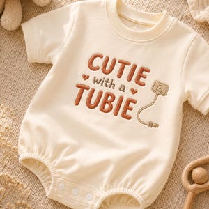 Könnte beinhalten: Ein cremefarbener kurzärmeliger Baby-Body mit dem gestickten Text "CUTIE with a TUBIE" in Braun, akzentuiert durch zwei kleine braune Herzen und eine gestickte Abbildung eines medizinischen Ernährungssonden-Anschlusses auf der rechten Seite.