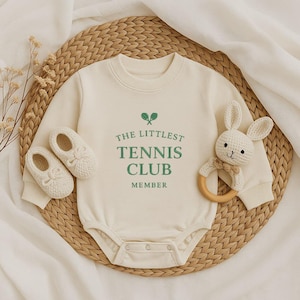 Op de afbeelding: Crèmekleurige babybody met de groene tekst "THE LITTLEST TENNIS CLUB MEMBER". De afbeelding bevat bijpassende babyschoentjes en een gehaakte konijnenrammelaar met een houten ring. De items zijn gerangschikt op een geweven mat.