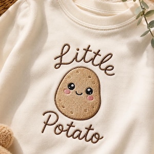 Pode incluir: Uma camisola de manga comprida cor de creme com um design bordado de uma batata sorridente com bochechas cor-de-rosa. O texto bordado em castanho acima e abaixo da batata diz "little" e "Potato" numa fonte de escrita cursiva.