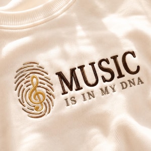 Può includere: Una felpa color crema con un ricamo che raffigura un'impronta digitale con una chiave di violino dorata al centro. A destra, il testo MUSIC IS IN MY DNA è ricamato con filo marrone scuro e grigio, con un girocollo a coste ben visibile.