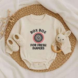 Peut inclure: Body de bébé crème avec le texte "BOX BOX FOR FRESH DIAPERS" en noir et rouge. Le body est accompagné de chaussures de bébé assorties et d'un hochet lapin blanc avec un anneau en bois.