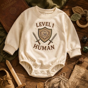 May include: An off-white long-sleeve bodysuit featuring an embroidered design on the chest. The embroidery includes the text "LEVEL 1 HUMAN" in brown, a central shield icon with a twenty-sided die, and two crossed swords positioned below the shield.