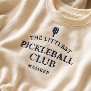 May include: A cream-colored crewneck sweatshirt featuring dark navy blue embroidery. The text reads THE LITTLEST PICKLEBALL CLUB MEMBER, accompanied by a small embroidered pickleball paddle icon centered above the lettering on the chest.