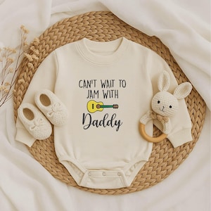 May include: Cream-colored baby onesie with the text "CAN'T WAIT TO JAM WITH Daddy" and a guitar graphic. The image includes matching baby shoes and a white bunny rattle with a wooden ring.