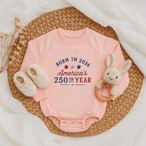 May include: A light pink baby onesie with the text "BORN IN 2026 America's 250th YEAR" in red and blue. The onesie is paired with white baby shoes and a stuffed bunny rattle with a wooden ring.