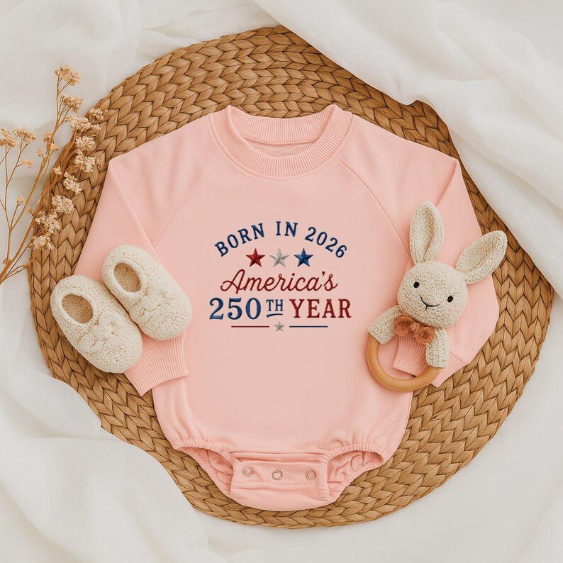 May include: A light pink baby onesie with the text "BORN IN 2026 America's 250th YEAR" in red and blue. The onesie is paired with white baby shoes and a stuffed bunny rattle with a wooden ring.
