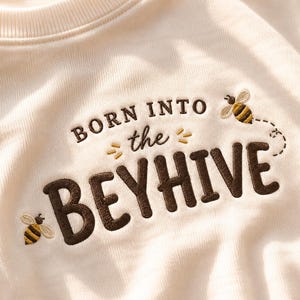 May include: A cream-colored sweatshirt featuring dark brown embroidered text that reads BORN INTO the BEYHIVE. The design includes two small embroidered bees with yellow and black stripes and white wings, with a dotted flight path trailing one bee.