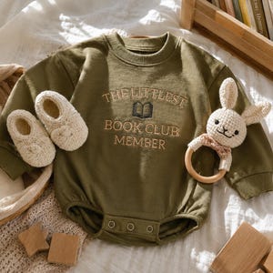 以下が含まれることがあります： オリーブグリーンの長袖ベビー用ボディスーツ。刺繍で「THE LITTLEST BOOK CLUB MEMBER」という文字と小さな本が描かれています。クルーネック、リブ編みの袖口、股下に3つのスナップボタンが付いています。クリーム色のニットブーティと、木製のリングが付いたかぎ針編みのウサギのガラガラが添えられています。