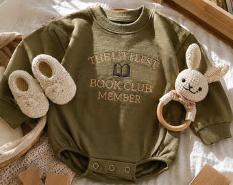May include: An olive green long-sleeve baby bodysuit featuring the embroidered text THE LITTLEST BOOK CLUB MEMBER and a small book icon. The garment includes a crew neckline, ribbed cuffs, and three snaps at the bottom. A pair of cream knit booties and a crochet rabbit rattle with a wooden ring are placed alongside.