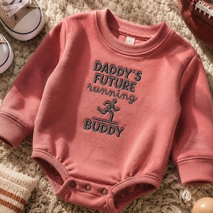 Può includere: Un body a maniche lunghe color rosa polvere con girocollo e polsini a coste. Al centro presenta un ricamo grigio scuro con il testo DADDY'S FUTURE running BUDDY e una piccola grafica raffigurante una persona che corre su una linea.