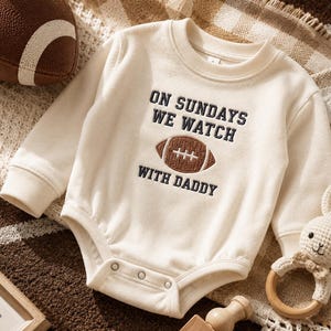 Pode incluir: Um body de bebé de manga comprida na cor creme com texto bordado em azul-marinho que diz ON SUNDAYS WE WATCH WITH DADDY, centrado em torno de um gráfico de uma bola de futebol americano castanha. A peça tem gola redonda e fechos de pressão.