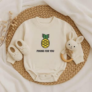 Op de afbeelding: Crème babybody met een gele en groene ananasafbeelding en de tekst "PINING FOR YOU". De afbeelding bevat ook crèmekleurige babyschoenen en een witte konijnenrammelaar met een houten ring.