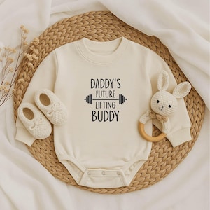 May include: Cream-colored baby bodysuit with the text "DADDY'S FUTURE LIFTING BUDDY" in black. The image includes a pair of white baby shoes and a white bunny rattle with a wooden ring. The items are arranged on a woven mat.