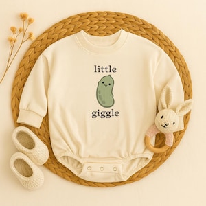 Könnte beinhalten: Cremefarbener Baby-Strampler mit dem Text "little giggle" und einer Cartoon-Gurke. Der Strampler wird mit einem kleinen Paar cremefarbener Babyschuhe und einem hölzernen Beißring in Hasenform kombiniert. Die Artikel sind auf einer gewebten Matte angeordnet.