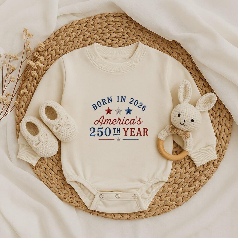 May include: Cream-colored baby onesie with the text "BORN IN 2026 America's 250th YEAR" in red and blue. The image includes a pair of white baby shoes and a white bunny-shaped rattle with a wooden ring.