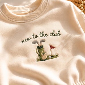 Könnte beinhalten: Ein cremefarbenes Sweatshirt mit Rundhalsausschnitt und einer Stickerei auf der Brust. Das Motiv zeigt eine Golftasche mit Schlägern, eine Fahne und einen Golfball auf einer grünen Fläche, ergänzt durch den Text "new to the club" in Dunkelgrün.