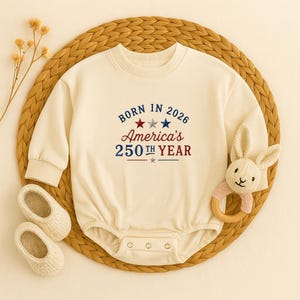 May include: Cream-colored baby bodysuit with long sleeves and the text "BORN IN 2026 America's 250th YEAR" in red and blue. The bodysuit is on a woven mat with a small pair of shoes and a bunny rattle.