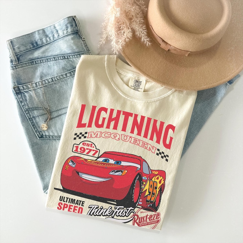 May include: A cream-colored t-shirt featuring a graphic of the red race car Lightning McQueen with the text Lightning McQueen, est. 1977, Ultimate Speed, Think Fast, and Rust-eze. The shirt is paired with light blue denim jeans, a tan hat, and a necklace.