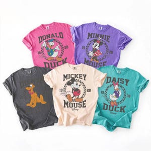 Op de afbeelding: Vijf t-shirts met korte mouwen in roze, paars, donkergrijs, cr&egrave;me en groenblauw. Elk shirt heeft een Disney-personage zoals Donald Duck, Minnie Mouse, Pluto, Mickey Mouse en Daisy Duck, met tekst en het jaartal 1928.