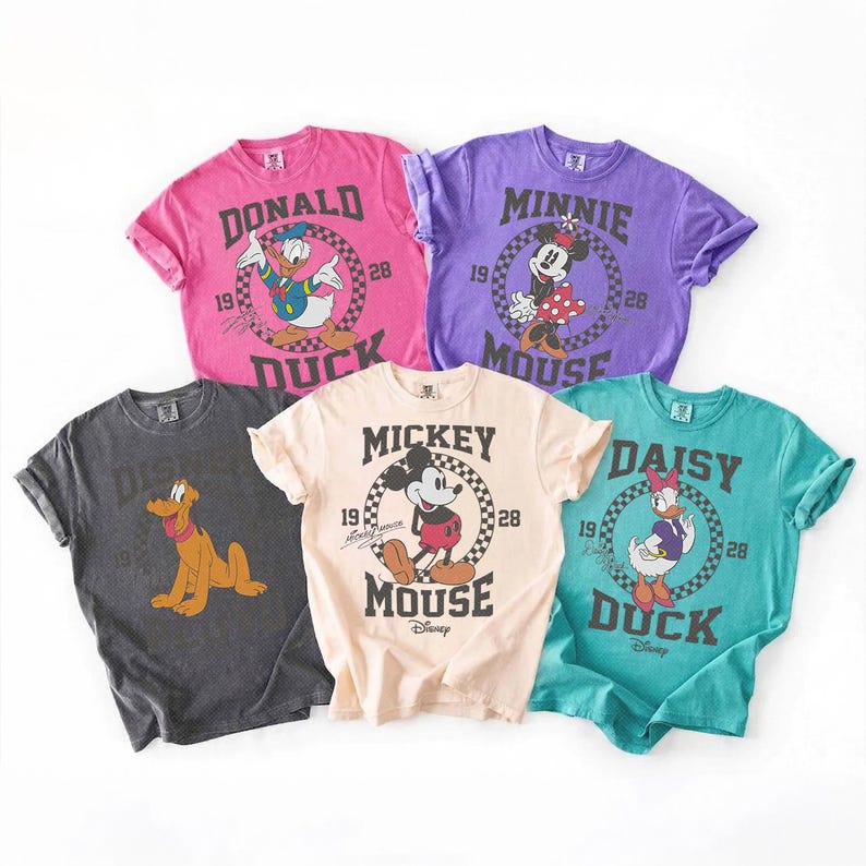 Op de afbeelding: Vijf t-shirts met korte mouwen in roze, paars, donkergrijs, cr&egrave;me en groenblauw. Elk shirt heeft een Disney-personage zoals Donald Duck, Minnie Mouse, Pluto, Mickey Mouse en Daisy Duck, met tekst en het jaartal 1928.