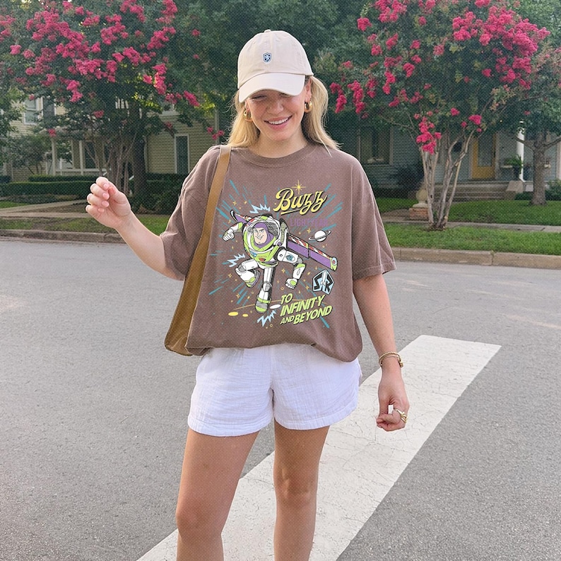 May include: A brown short-sleeve t-shirt featuring a graphic of Buzz Lightyear with the text Buzz To Infinity And Beyond, paired with white shorts, a tan baseball cap, and a brown shoulder bag. The graphic includes stars and planets.