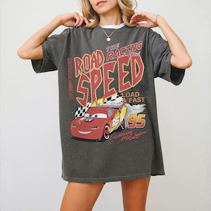 May include: A charcoal gray short-sleeve t-shirt featuring a graphic print of a red race car with the text THE ROAD RACING CLUB SPEED, LOAD & FAST, 95, SUPERIOR PERFORMANCE, and Lightning McQueen. The design includes flames and a checkered flag.
