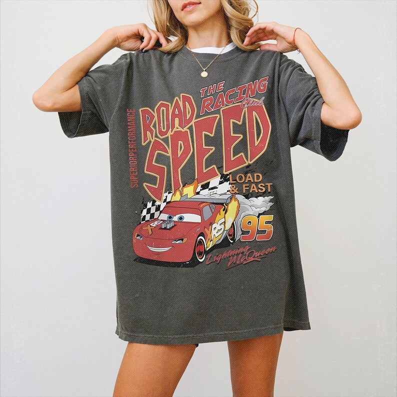 May include: A charcoal gray short-sleeve t-shirt featuring a graphic print of a red race car with the text THE ROAD RACING CLUB SPEED, LOAD & FAST, 95, SUPERIOR PERFORMANCE, and Lightning McQueen. The design includes flames and a checkered flag.