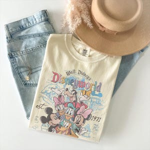 May include: A cream-colored short-sleeve t-shirt featuring a graphic print of Mickey Mouse, Minnie Mouse, Donald Duck, Daisy Duck, and Goofy. The text reads Walt Disney's Disneyworld est. 1971. The shirt is paired with light blue denim jeans and a tan hat.