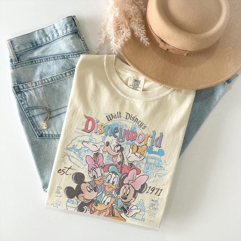 May include: A cream-colored short-sleeve t-shirt featuring a graphic print of Mickey Mouse, Minnie Mouse, Donald Duck, Daisy Duck, and Goofy. The text reads Walt Disney's Disneyworld est. 1971. The shirt is paired with light blue denim jeans and a tan hat.