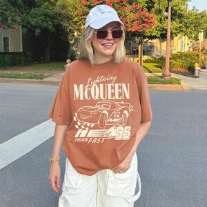 May include: A rust-colored short-sleeve t-shirt featuring a graphic of the character Lightning McQueen with the text Lightning MCQUEEN and THINK FAST, paired with a white baseball cap and white cargo pants with large pockets.