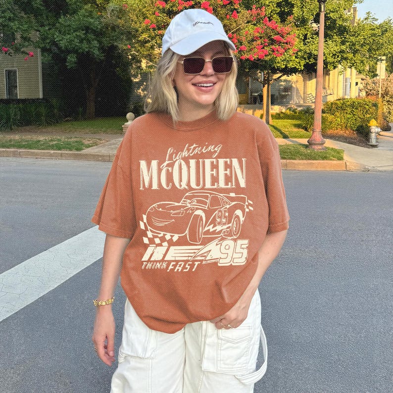 May include: A rust-colored short-sleeve t-shirt featuring a graphic of the character Lightning McQueen with the text Lightning MCQUEEN and THINK FAST, paired with a white baseball cap and white cargo pants with large pockets.