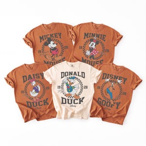 Op de afbeelding: Vijf t-shirts met korte mouwen met Disney-figuren. Vier shirts zijn roestkleurig en &eacute;&eacute;n is cr&egrave;me. Elk shirt toont een cirkelvormige dambordafbeelding met de naam van het personage, het jaar 1928 en een illustratie van Mickey, Minnie, Daisy, Donald of Goofy.