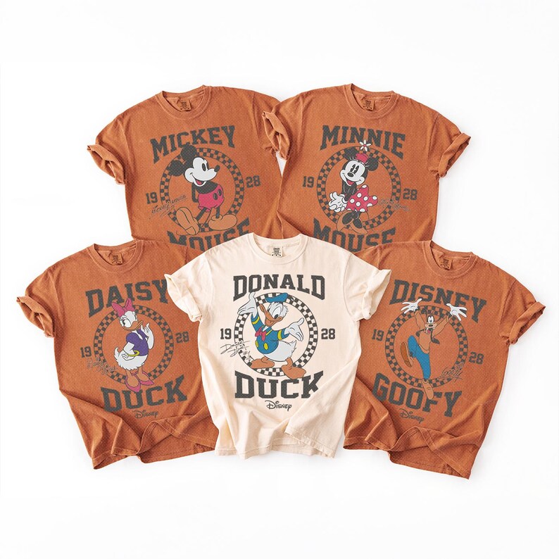 Op de afbeelding: Vijf t-shirts met korte mouwen met Disney-figuren. Vier shirts zijn roestkleurig en &eacute;&eacute;n is cr&egrave;me. Elk shirt toont een cirkelvormige dambordafbeelding met de naam van het personage, het jaar 1928 en een illustratie van Mickey, Minnie, Daisy, Donald of Goofy.