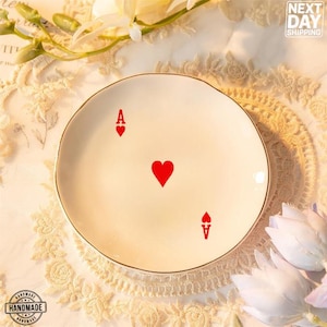 May include: A white ceramic trinket dish with a gold rim. The dish features a red ace of hearts playing card design.