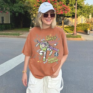 May include: A rust-colored short-sleeve t-shirt featuring a graphic of Buzz Lightyear with the text "Buzz Lightyear" and "To Infinity and Beyond" in a stylized font, accompanied by a white baseball cap and white cargo pants.