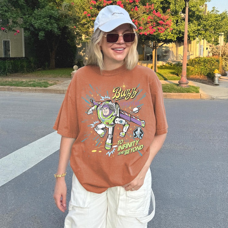 May include: A rust-colored short-sleeve t-shirt featuring a graphic of Buzz Lightyear with the text "Buzz Lightyear" and "To Infinity and Beyond" in a stylized font, accompanied by a white baseball cap and white cargo pants.